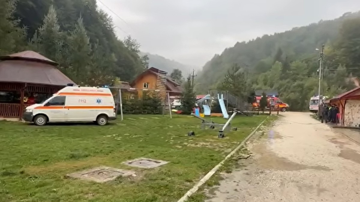 O ambulanță și alte vehicule de intervenție staționate în curtea unei cabane, înconjurată de munți