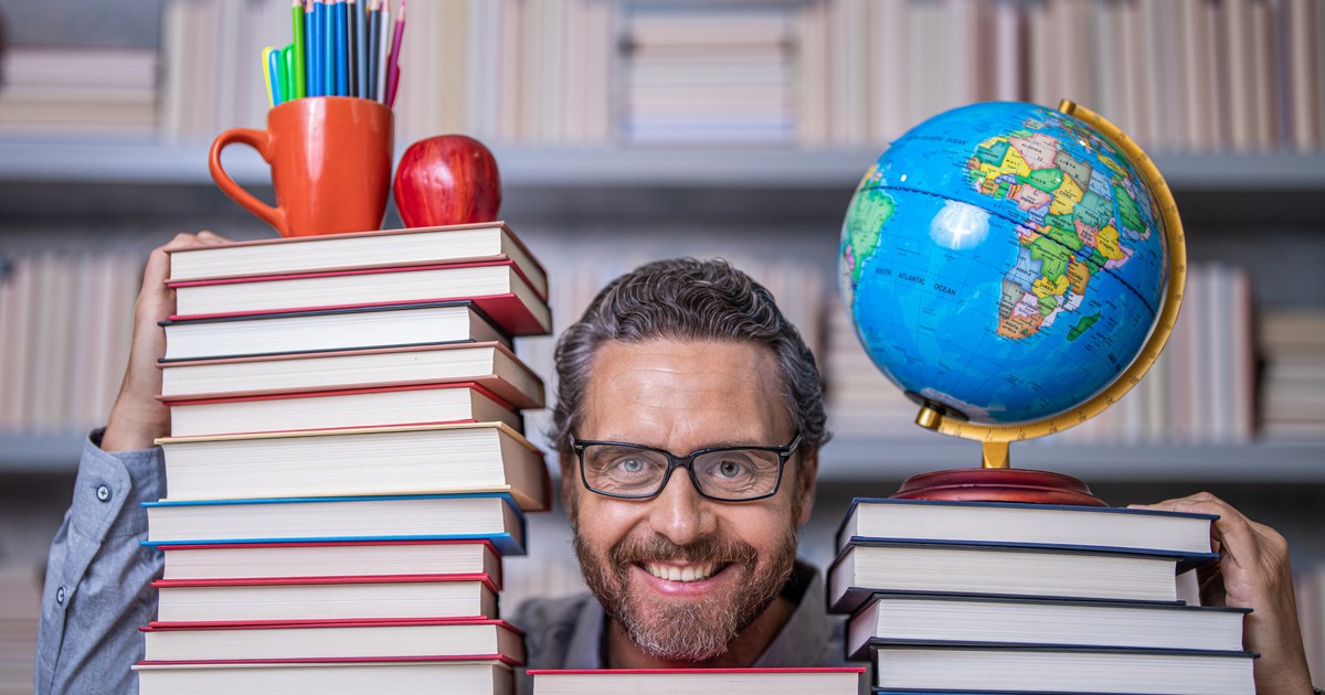 Profesor cu ochelari înconjurat de mai multe teancuri de cărți