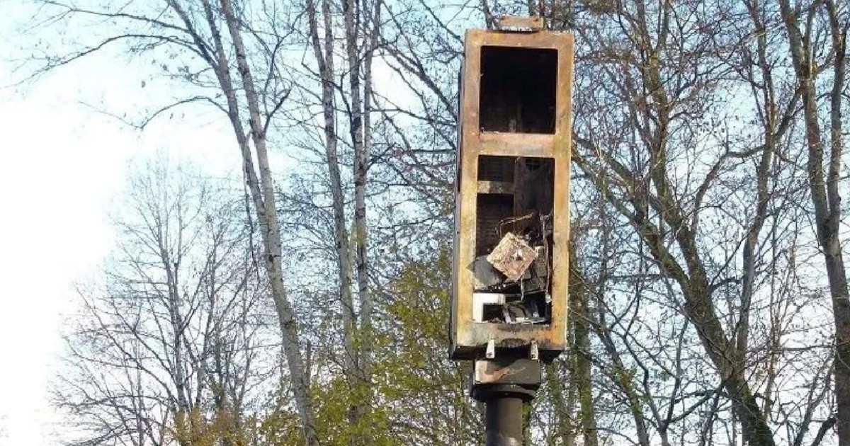 Un radar de viteză distrus, montat pe un stâlp, lângă copaci fără frunze, sub un cer albastru.
