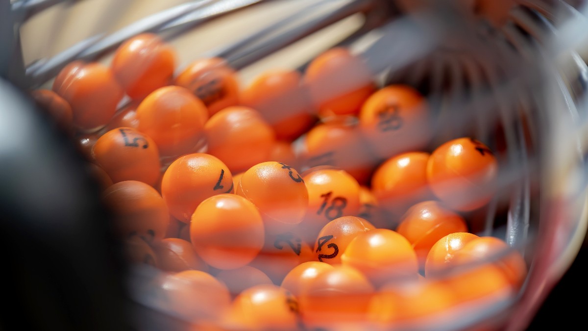 În imagine se văd bile portocalii de loto, numerotate, aflate într-un dispozitiv transparent folosit la extragere. Fotografia surprinde momentul amestecării bilelor, sugerând emoția și așteptarea specifică unui joc de noroc.