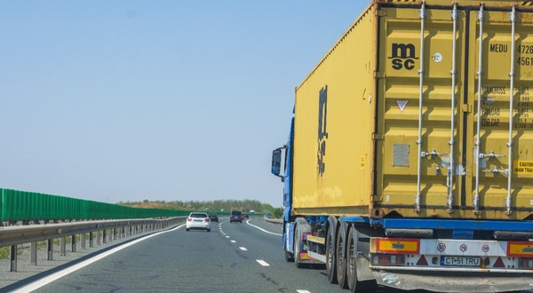 Imaginea surprinde un camion albastru ce transportă un container galben, rulând pe o autostradă alături de alte autoturisme, sub un cer senin.