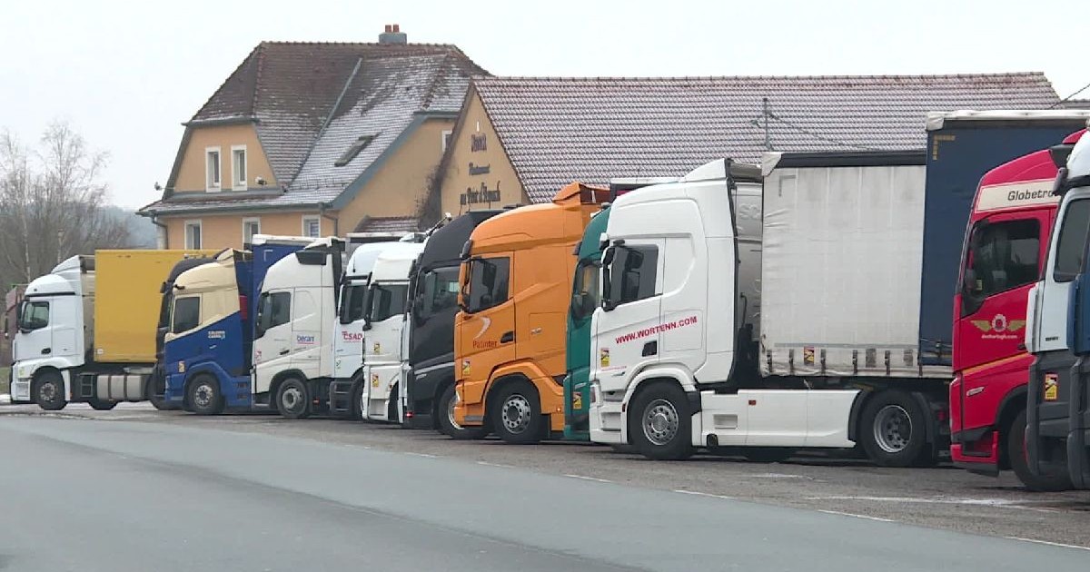 Un șir de camioane mari, inclusiv unul portocaliu, parcate pe un drum în fața unor clădiri, copaci în fundal.