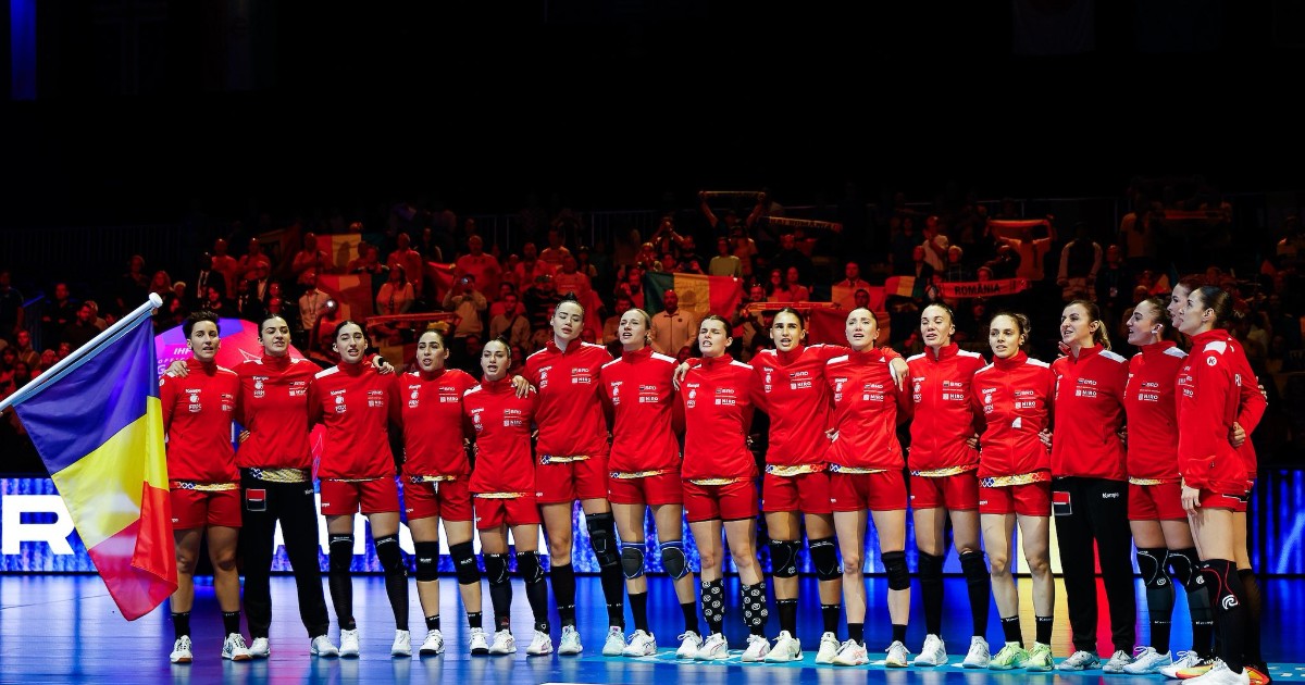 În imagine apare echipa națională de handbal feminin a României, aliniată pe teren înainte de startul meciului, îmbrăcată în echipament roșu oficial. Jucătoarele stau una lângă alta, ținându-se de umeri, în timp ce un drapel tricolor este fluturat în fața lor, pe fundal fiind vizibili suporterii români din tribune.