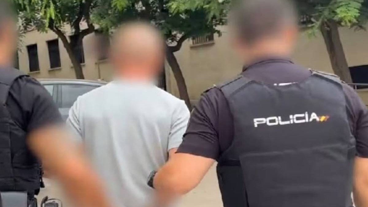 Doi polițiști spanioli escortează un bărbat încătușat, îmbrăcat într-un tricou deschis la culoare. Imaginea este realizată din spate, pe o stradă, iar fețele celor trei persoane sunt blurate. În colțul din stânga sus apare sigla Poliției Naționale din Spania.