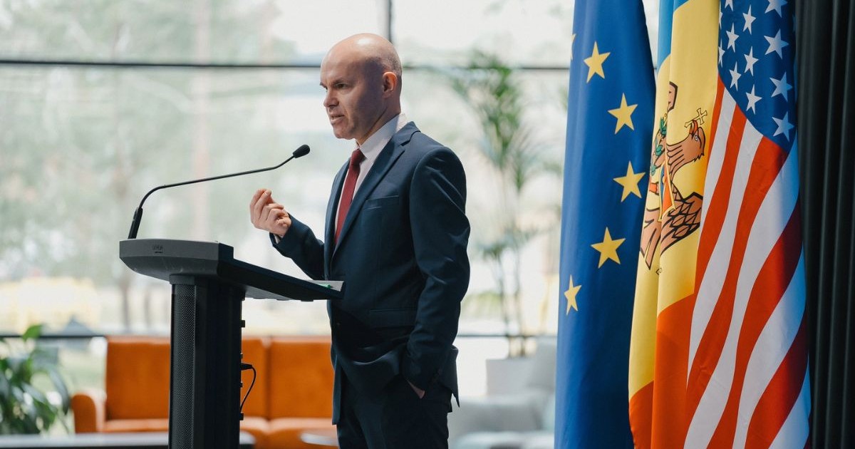 Daniel Funeriu in costum la un pupitru cu microfon avand in spate steagurile UE, Romaniei si SUA
