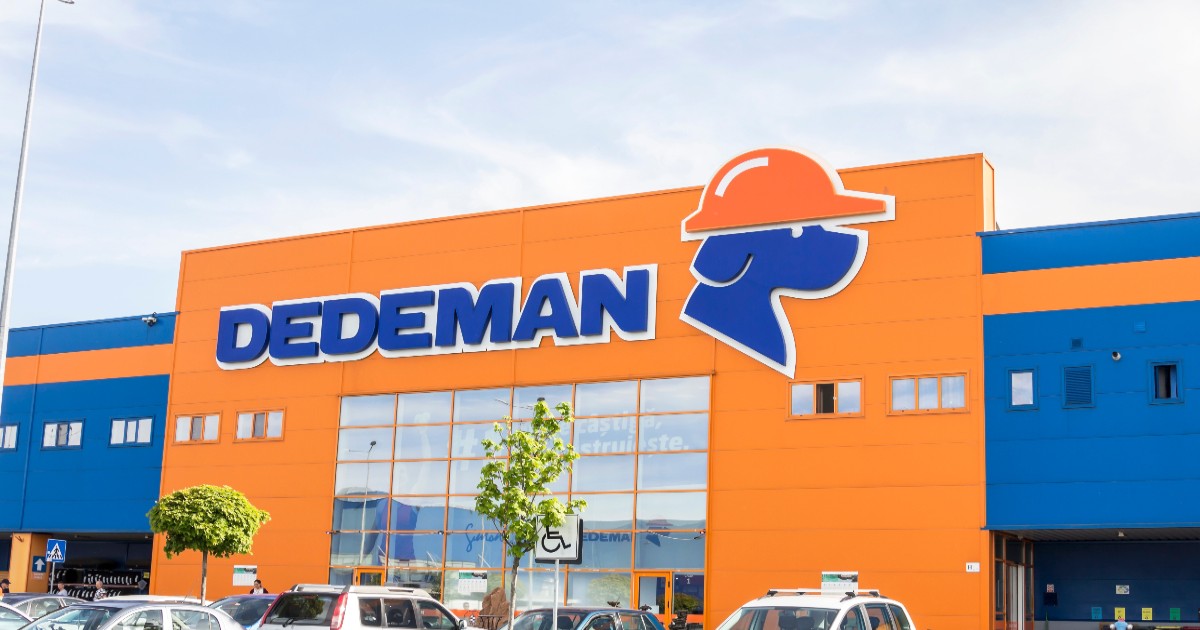 Imaginea surprinde un magazin Dedeman, cu fațada sa distinctivă în culorile portocaliu și albastru, pe care este afișat logo-ul companiei. În fața clădirii se află o parcare cu mai multe autoturisme, semn că magazinul este deschis și frecventat de clienți.