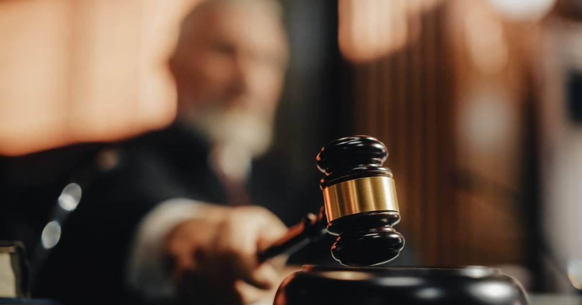 Un judecător care stă pe scaun în sala de instanță, poartă o robă neagră și ridică ciocănelul, în timp ce urmează să pronunțe un verdict