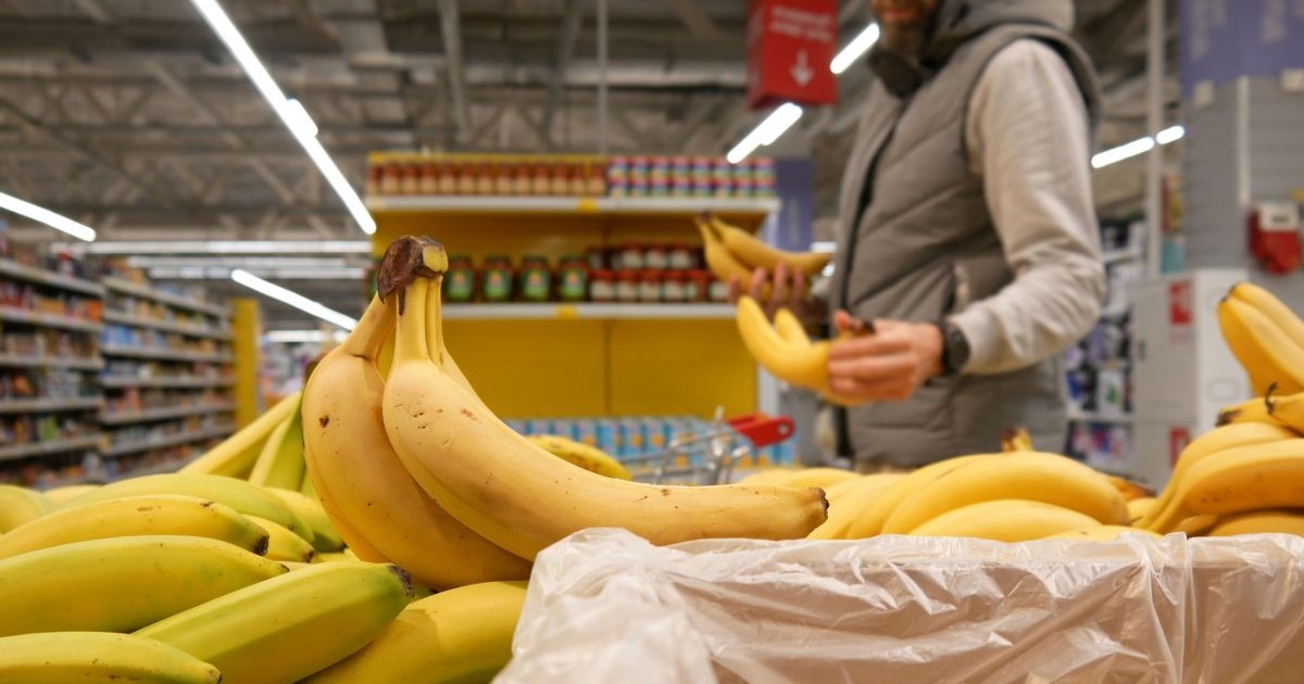 Banane coapte în cutii, pe raftul unui supermarket, și un bărbat care ține o banană în mână, pe fundal