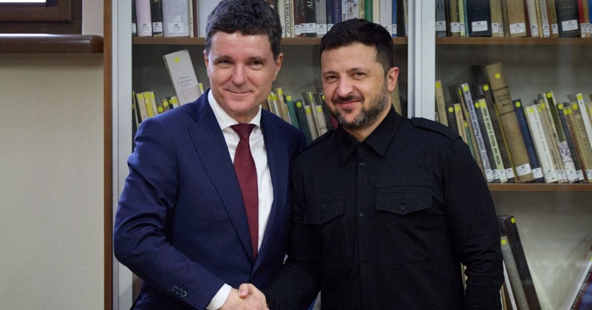 Nicușor Dan, îmbrăcat în costum albastru, cămașă albă și cravată roșie și Volodimir Zelenski, când dau noroc, îmbrăcat cu o bluză neagră, iar pe fundal o bibliotecă plină cu cărți