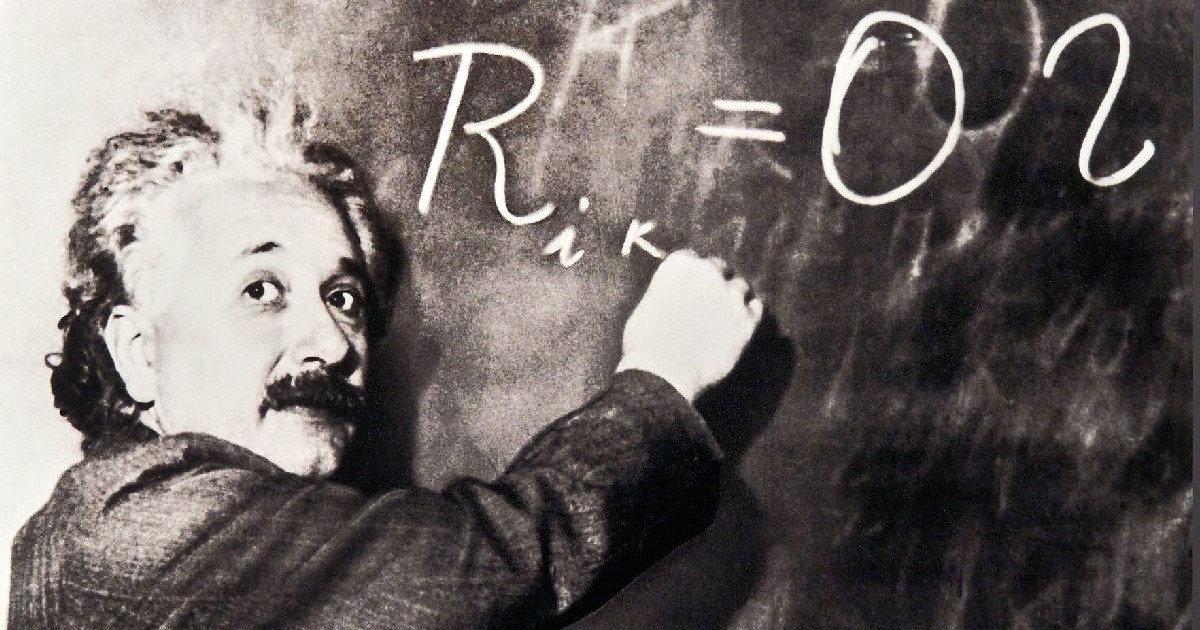 O fotografie alb-negru cu Albert Einstein, îmbrăcat în costum, scriind o ecuație pe tablă