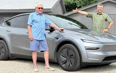 Un bărbat de 98 de ani, cu camasa albastra si pantaloni scurti albastri, langa o masina Tesla gri, pe partea opusa, un bărbat cu camasa verde