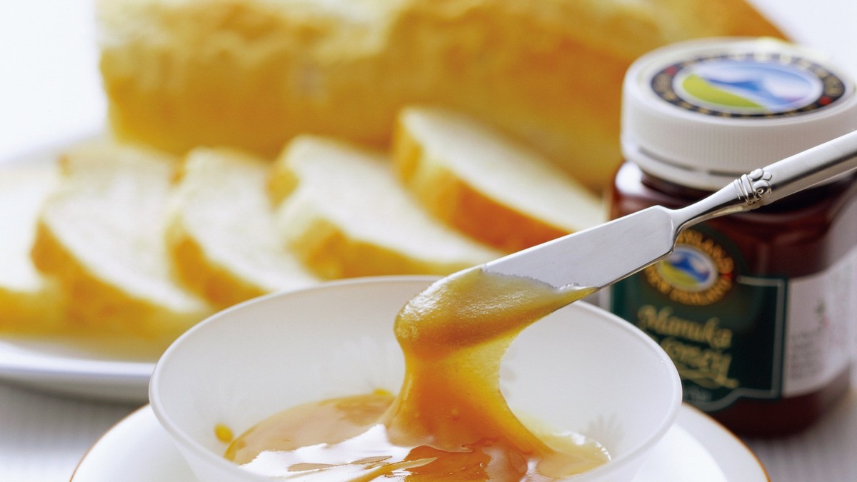 Oferta ManukaShop.ro de azi - Un bol cu miere pe o farfurie, lângă un borcan de miere de Manuka şi mai multe felii de pâine