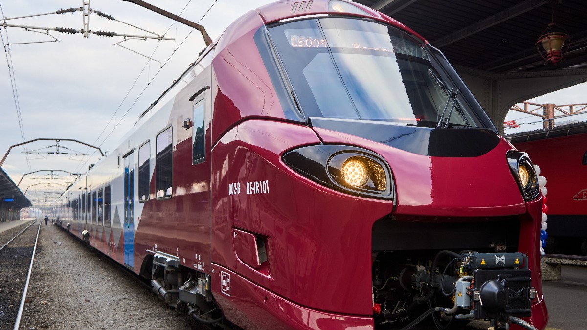 Fotografia surprinde un tren modern, de culoare roșie-grena, cu accente albe și gri, aflat într-o gară. Partea din față a locomotivei este rotunjită și are două faruri rotunde, cu lumina aprinsă. Pe laterale se pot observa vagoanele. Imaginea este realizată din unghiul șinelor, dând o senzație de viteză și dinamism.