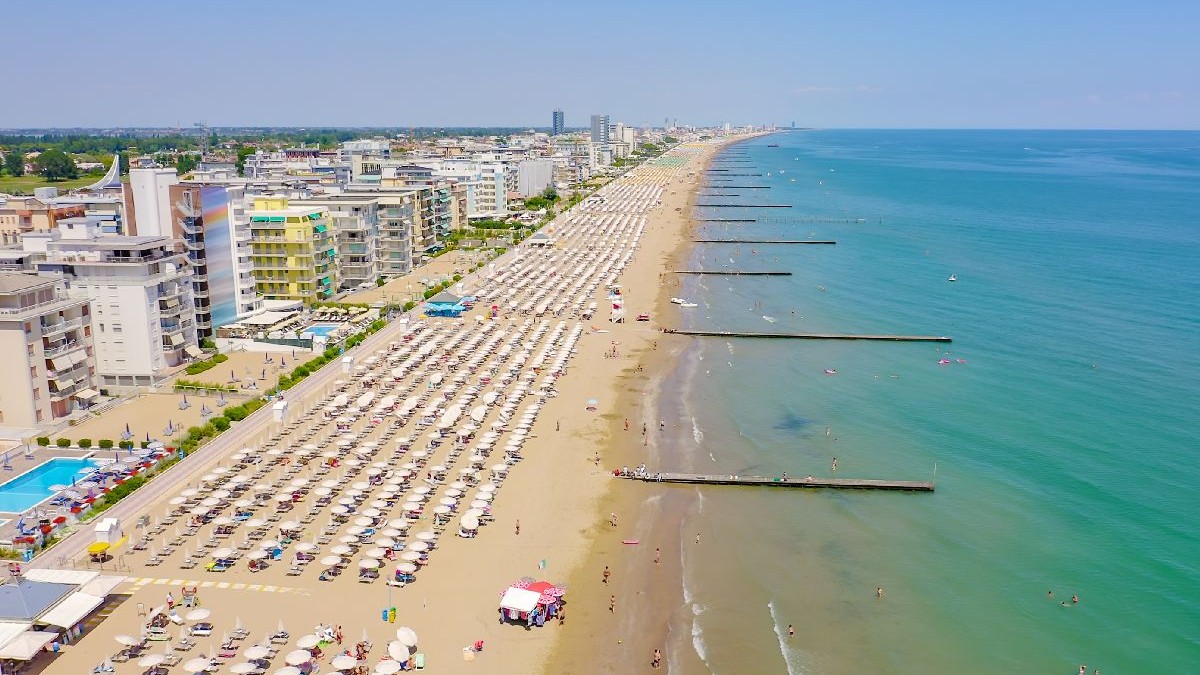 Fotografie aeriană a unei plaje, cu hoteluri, rânduri de umbrele albe și șezlonguri și apă albastră cristalină.