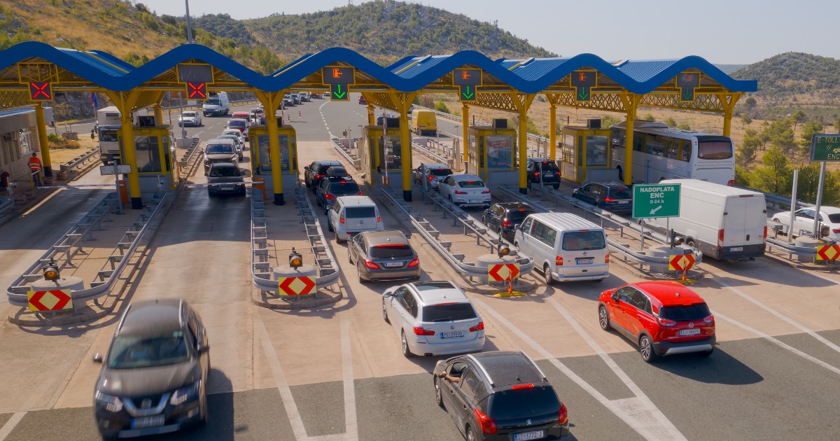 Imaginea prezintă o stație de taxare pe o autostradă, unde numeroase vehicule se află la coadă pentru a trece prin cabinele de plată.