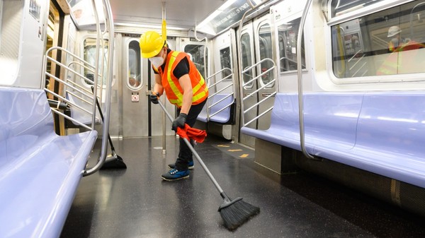 Un angajat la curățenie purtând cască de protecție, vestă reflectorizantă și mască mătură podeaua în interiorul unui vagon de metrou.   