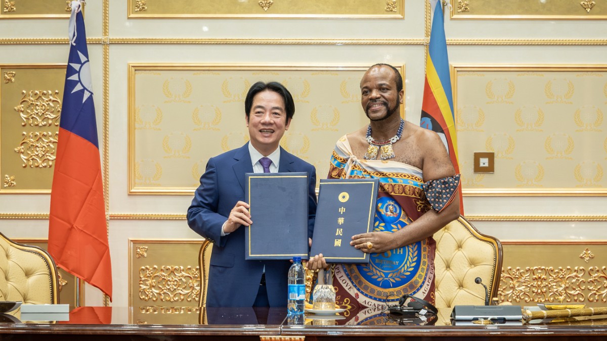 Președintele Taiwanului, Lai Ching-te, și Regele Mswati al III-lea al Eswatini-ului stau alături și prezintă dosare oficiale semnate. Cei doi oficiali zâmbesc în fața steagurilor celor două țări, într-o sală de protocol decorată elegant în nuanțe aurii.