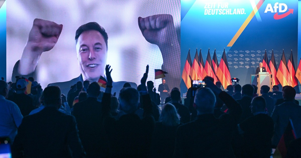 elon musk in timp ce participa online la un miting in germania. imaginea lui este proiectata pe un ecran mare iar in fata este publicul care aplauda