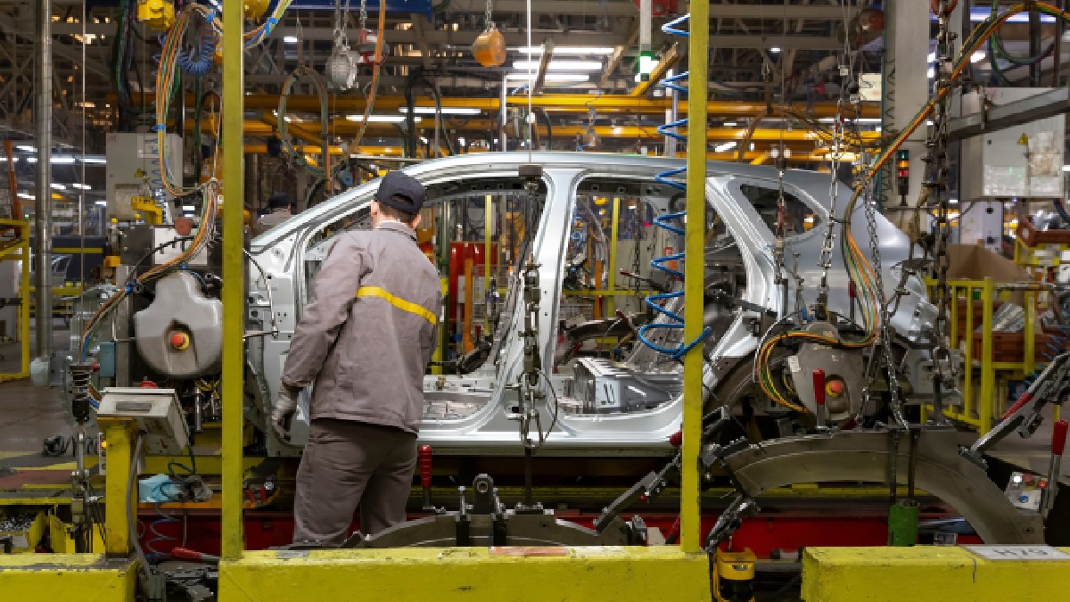 Un muncitor dintr-o fabrică auto monitorizând caroseria unui vehicul aflată pe o linie de asamblare, înconjurată de diverse utilaje și echipamente industriale.