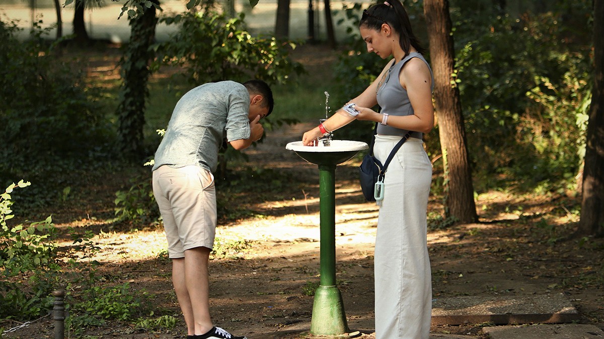 Două persoane lîngă o țâșnitoare de apă dintr-un parc al Bucureștiului se spală pe față și pe mâini.