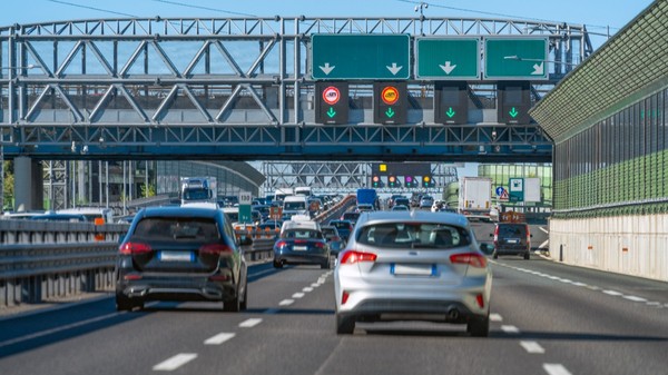 Trafic intens de vehicule pe o autostradă aglomerată, cu mai multe benzi circulabile și panouri electronice de semnalizare montate pe o structură metalică suspendată.