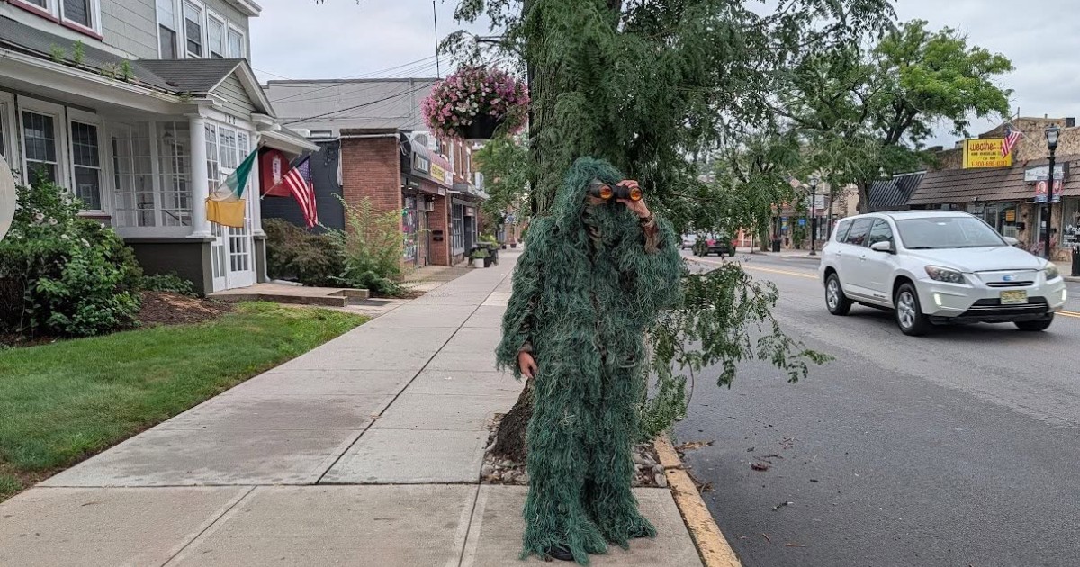 Imaginea surprinde o persoană îmbrăcată într-un costum de camuflaj verde, asemănător unui copac sau unui arbust, care privește printr-un binoclu pe o stradă dintr-o zonă urbană. Costumul acoperă aproape în întregime corpul și capul, iar în jur se observă copaci, magazine și autoturisme parcate.