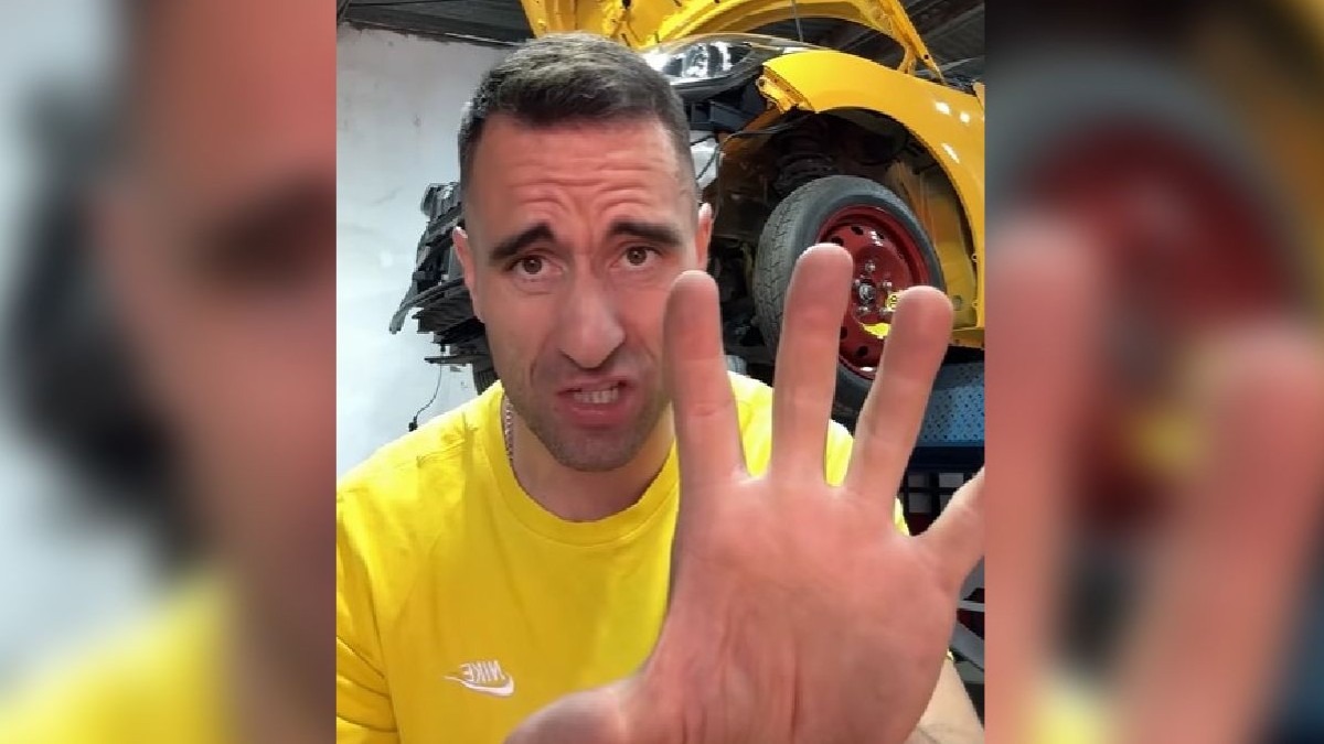 Imaginea arată un bărbat îmbrăcat într-o bluză galbenă care arată cinci degete spre cameră, având în fundal o mașină galbenă ridicată pe o platformă într-un atelier auto.