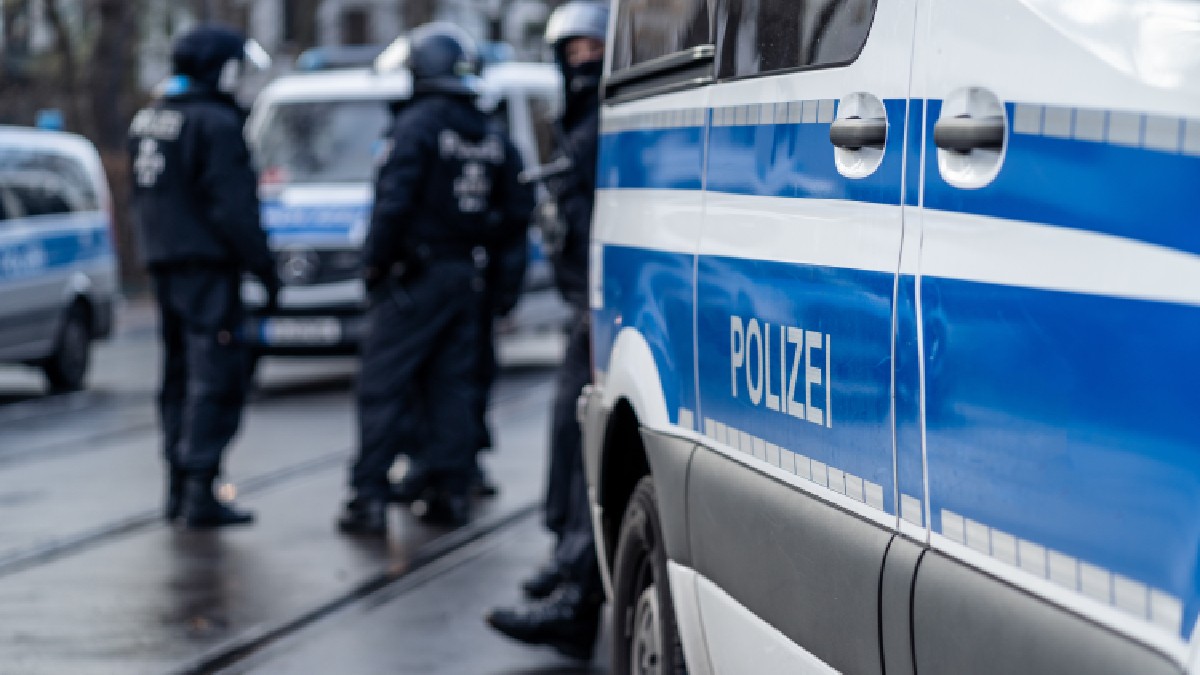 Imaginea prezintă o scenă de pe o stradă, unde se află mai multe vehicule și ofițeri de poliție germani. În prim-plan, se vede clar o porțiune a unei furgonete de poliție, de culoare albă și albastră, pe care este inscripționat cuvântul "POLIZEI". În spatele furgonetei din prim-plan, se pot distinge alți ofițeri de poliție, îmbrăcați în uniforme de culoare închisă și purtând căști. De asemenea, se văd și alte vehicule ale poliției, parțial vizibile în fundal. Străzile par umede, sugerând că ar fi putut ploua recent.
