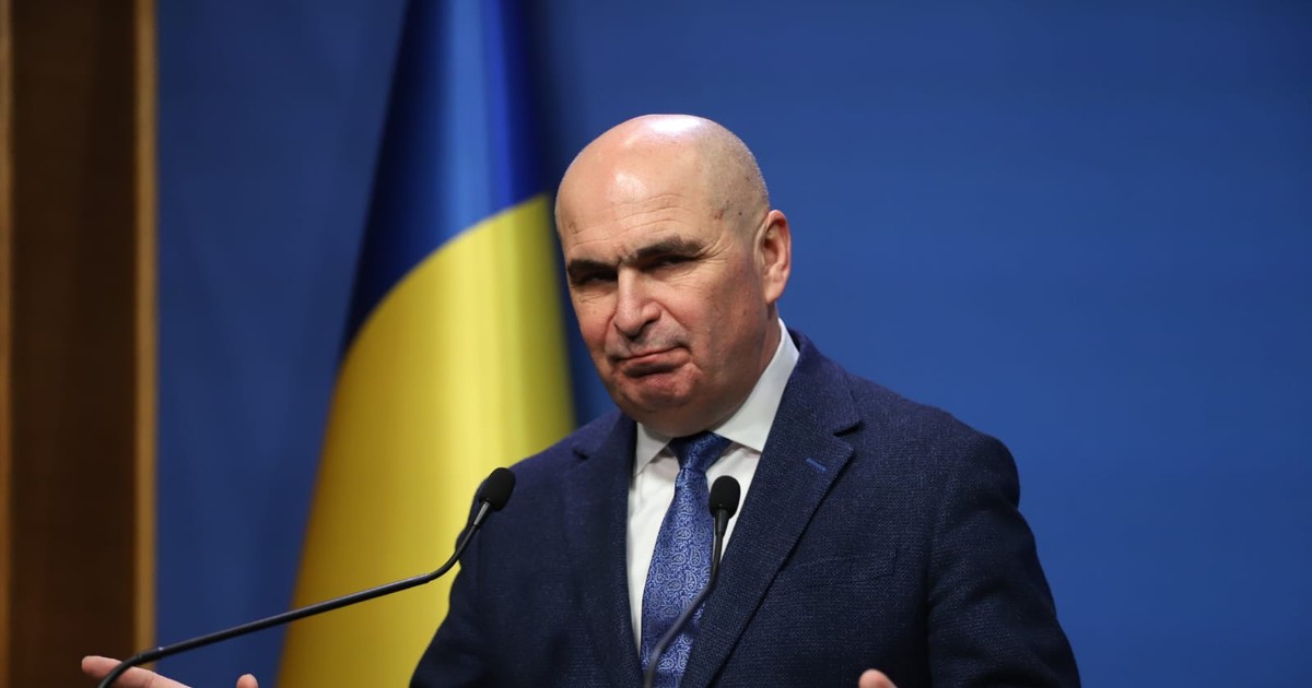Ilie Bolojan in conferinta de presa, gesticuleaza larg cu mainile. Ilie Bolojan poarta un costum albastru deschis, o camasa alba si cravata albastru deschis. In spate se vede un drapel tricolor