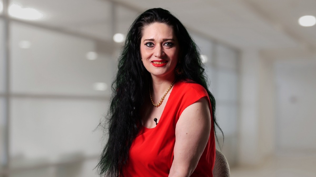 Dr. Sorina Ispas, îmbrăcată cu o bluză roșie, pe fundal blurat, cu părul lung negru, în timp ce stă pe un scaun