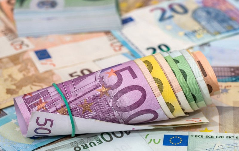 Sul de bancnote euro cu valori diferite legate cu un elastic, așezat pe un fundal acoperit de numeroase alte bancnote euro împrăștiate