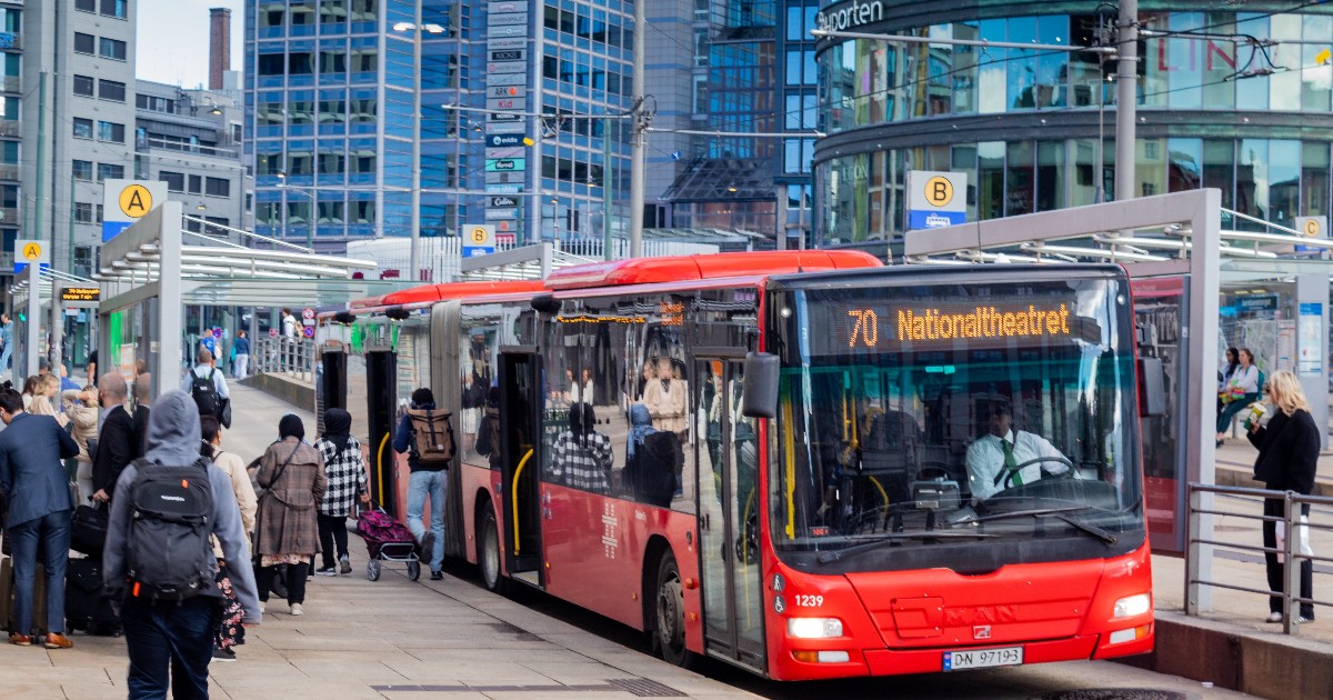 Imaginea surprinde un autobuz articulat de culoare roșie, oprit într-o stație aglomerată, cu uși deschise pentru îmbarcarea și debarcarea pasagerilor. Autobuzul poartă numărul liniei 70 și destinația "Nationaltheatret" afișată pe ecran, în timp ce pe fundal se văd clădiri moderne din sticlă și o zonă urbană dens populată.