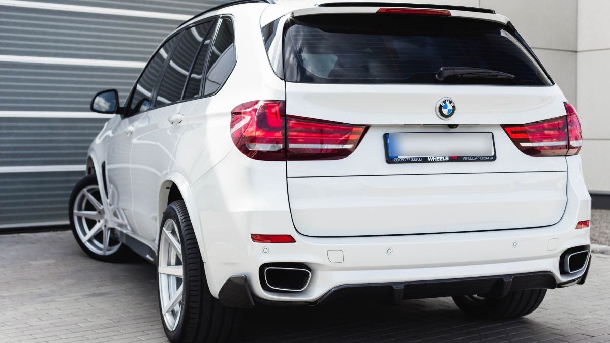 Fotografie cu un SUV BMW X5 alb, cu un design sportiv, parcat pe o suprafață pavată în fața unei uși de garaj gri. Mașina are geamuri fumurii, două țevi de eșapament și jante din aliaj.