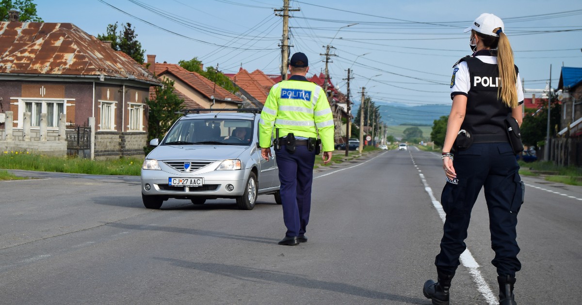 Imaginea prezintă doi polițiști români aflați pe carosabil într-o zonă rurală, dintre care unul oprește pentru control o mașină argintie.