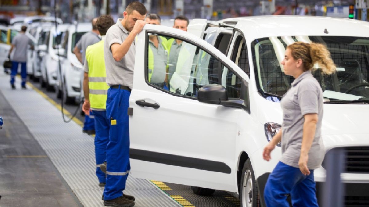 Imaginea prezintă o linie de asamblare a mașinilor, cu muncitori îmbrăcați în salopete albastre și veste de siguranță, vehicule albe și o femeie blondă îmbrăcată într-o cămașă gri și pantaloni albaștri