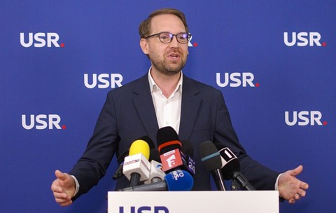Dominic Fritz îmbrăcat într-un sacou negru și o cămașă albă, vorbind în fața unui pupitru cu sigla USR pe care se află mai multe microfoane, cu mâinile larg deschise