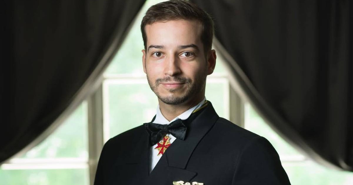 Jonas Lauwiner pozează într-o uniformă neagră elegantă, cu papion și decorații pe piept, în fața unei ferestre acoperite parțial de draperii închise la culoare. Bărbatul privește spre cameră și zâmbește discret.