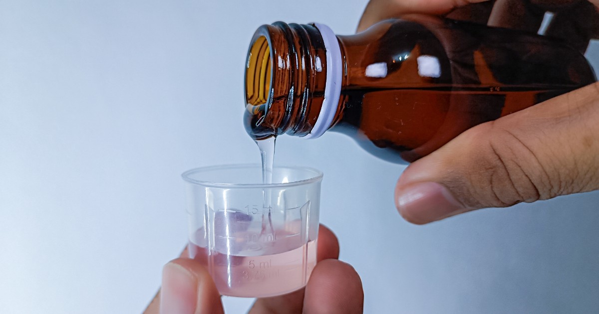 Imaginea, un prim-plan realizat în lumină puternică, surprinde momentul în care un lichid transparent este turnat dintr-o sticlă maro cu dop într-o cupă de măsurare din plastic. Recipientul de măsurare conține deja o cantitate mică de lichid de culoare roz, iar pe el sunt marcate gradări în mililitri.