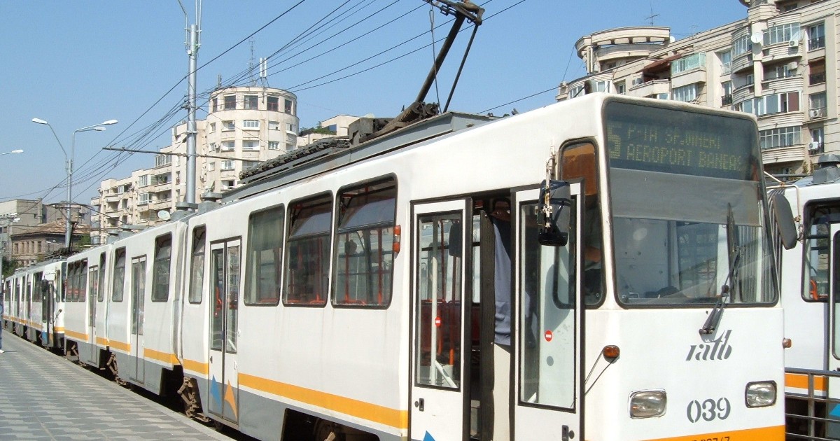 Imaginea prezintă un tramvai de culoare albă, cu bandă portocalie-galbenă, oprit într-o stație aglomerată din București. Pe afișajul frontal este indicat clar Traseul 5, cu destinația "P-ȚA SF. GHEORGHE / AEROPORT BĂNEASA", având în fundal clădiri de locuințe.