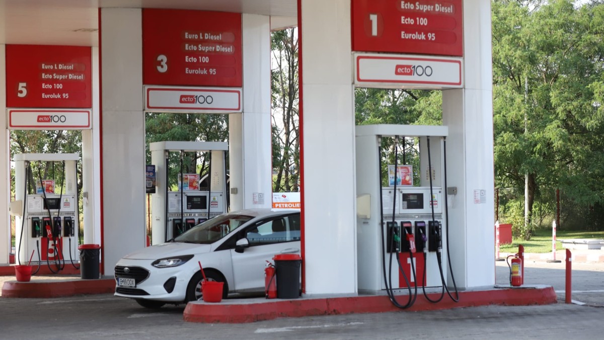 Imaginea prezintă o mașină albă parcată lângă pompele de alimentare ale unei stații de carburanți Lukoil.