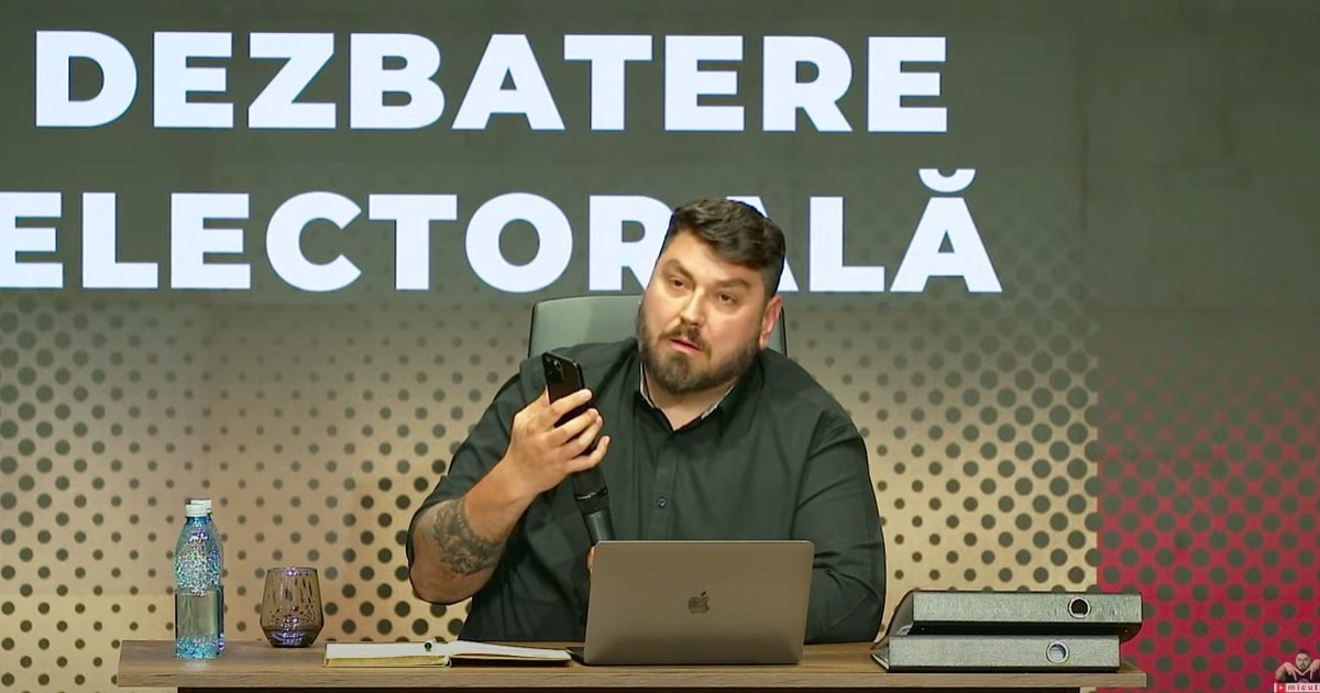 Micutzu alais Cosmin Nedelcu, in studio, la birou, cu un telefon in mana si un laptop in fata