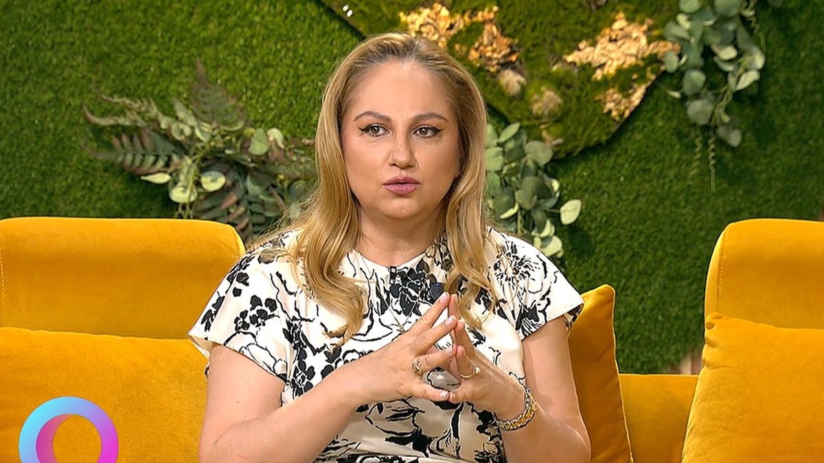 Astrologul Cristina Demetrescu, îmbrăcată cu o bluză alb cu negru, stă pe o canapea portocalie la o emisiune TV și are în spate un fundal verde
