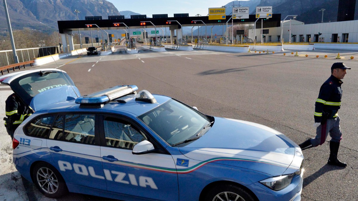 O mașină a poliției italiene este staționată lângă o stație de taxare de pe autostradă, în timp ce un ofițer patrulează zona în apropierea cabinelor de control.