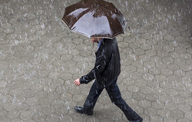 Un bărbat îmbrăcat într-o geacă neagră și pantaloni închiși la culoare, mergând pe un trotuar pavat în timp ce ține o umbrelă maro deasupra capului, pe o ploaie torențială.