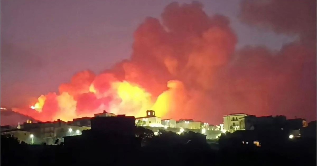 o așezare umană pe timp de noapte, profilată pe fundalul unui incendiu de proporții. În partea inferioară a imaginii se conturează siluetele unor clădiri de diferite dimensiuni, așezate pe un teren colinar, printre care se observă surse punctiforme de iluminat stradal. Imediat în spatele acestor construcții se ridică volume masive și dense de fum, care domină restul cadrului. Baza acestor formațiuni de fum este puternic luminată de flăcările intense ale focului care mistuie relieful din vecinătatea caselor, creând un contrast evident cu clădirile din prim-plan aflate în penumbră.