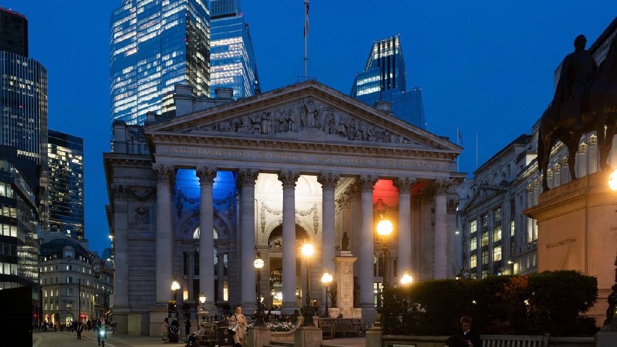 Royal Stock Exchange din Londra, iluminat în culorile drapelului francez, se profilează în fața zgârie-norilor moderni. Oamenii trec pe lângă el la apusul soarelui.