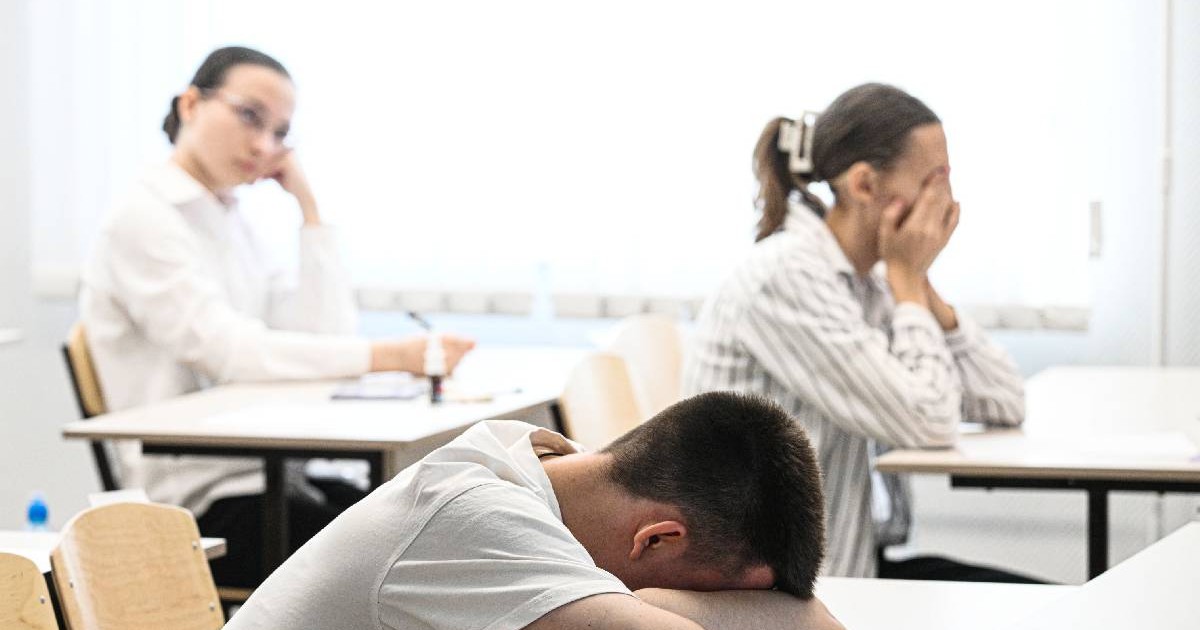 În imagine un elev stă cu capul sprijinit de mâini, pe bancă, o elevă își ține mâinile pe față dezamăgită iar o altă elevă privește spre partea din față a sălii de examen așteptând