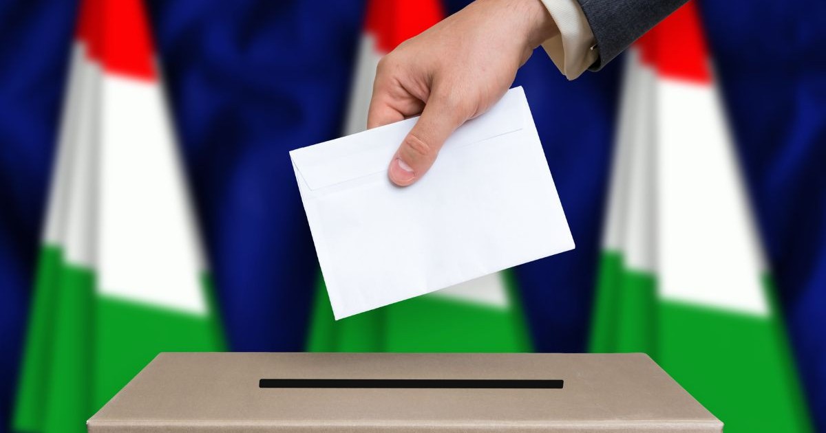 mana unui bărbat în sacou gri, sub care e vizibilă manșeta unei cămăși albe, introducând un vot într-o urnă, cu steagul ungariei pe fundal - alegeri parlamentare ungaria 12 aprilie 2026
