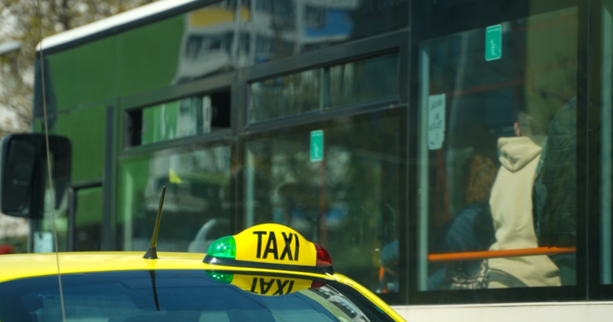 Imaginea surprinde partea din spate a unui taxi galben, specific Bucureștiului, cu semnalizarea luminoasă „TAXI” montată pe plafon. Lângă vehicul se vede un autobuz în care se află mai mulți pasageri. Scena este tipică pentru traficul urban, redând un moment obișnuit din transportul public al capitalei.
