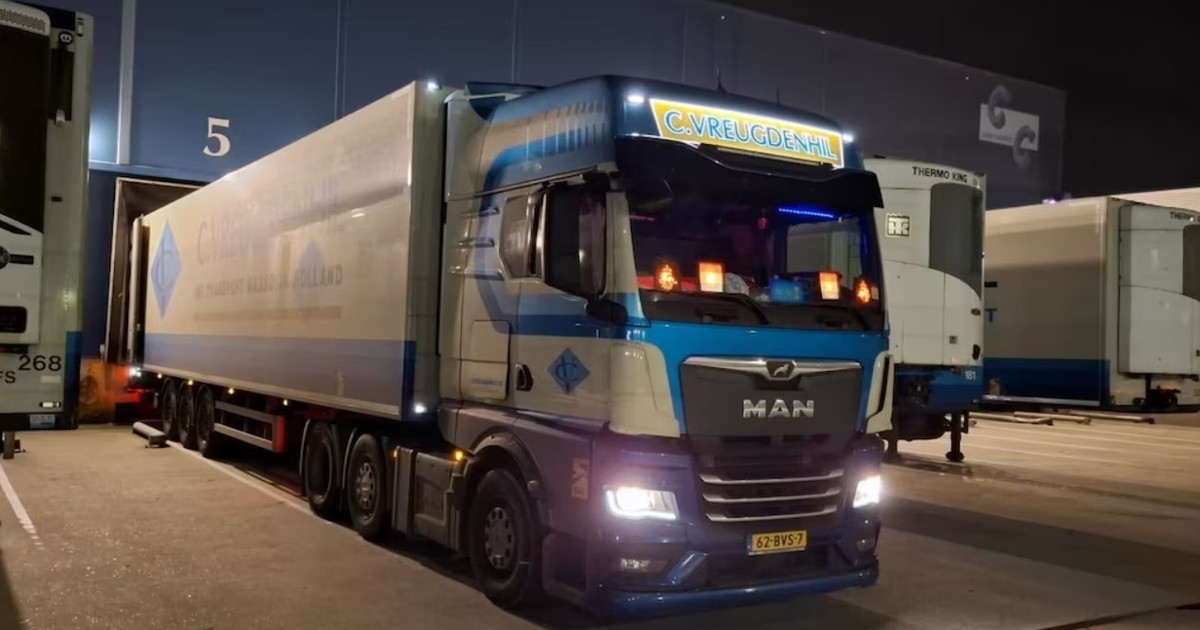 Fotografie surprinsă noaptea cu un camion MAN albastru parcat. Farurile și stopurile camionului sunt aprinse. Fundalul include un depozit slab iluminat.